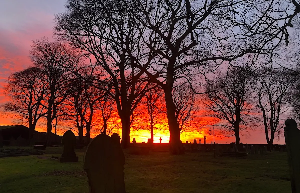 A fiery sunset behind the trees and headstones of St Lucius' churchyard.